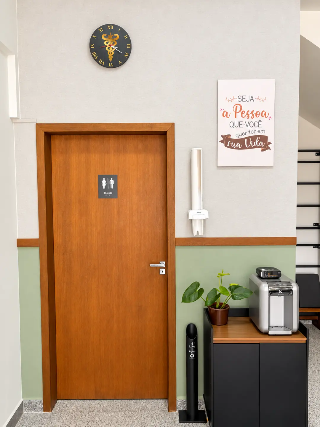 Área interna da clínica com porta do sanitário, bebedouro, planta decorativa e lixeira de reciclagem.