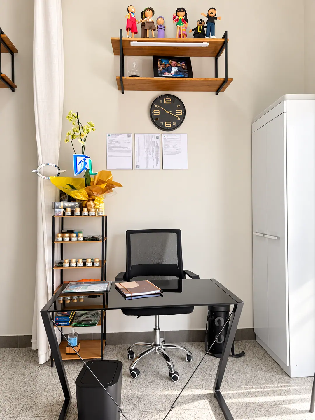 Consultório com mesa de atendimento, estantes com materiais, quadro de horários e armário branco.