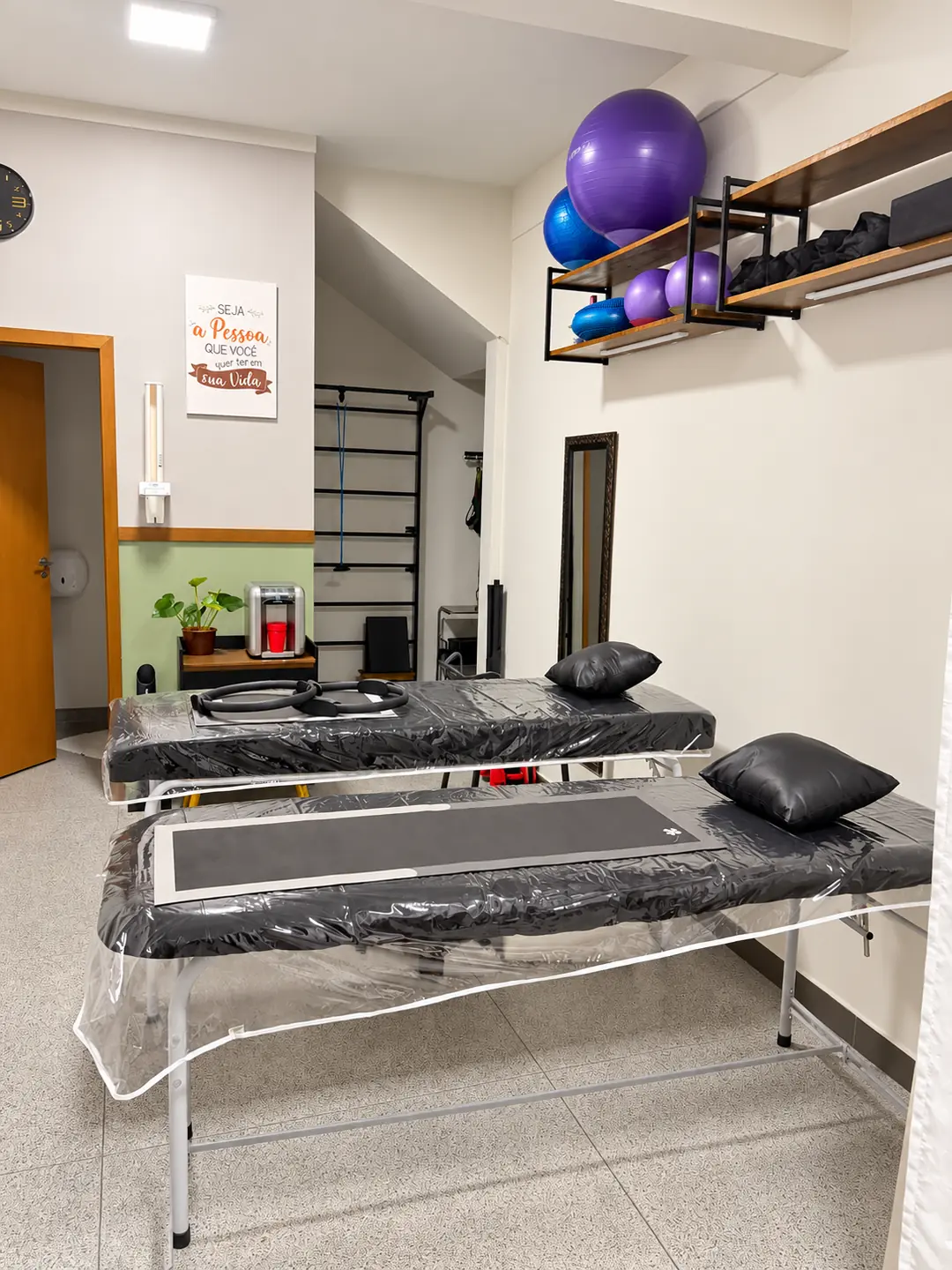 Sala de atendimento com duas macas de tratamento, bolas de exercício nas prateleiras e espaldeira ao fundo.